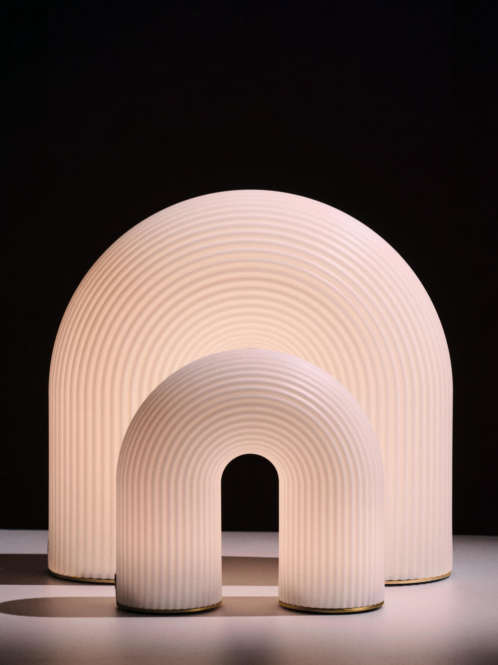 Two ribbed arch-shaped lamps in white, lit, in two sizes against a dark background