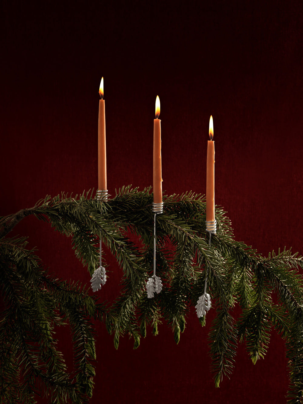 Three candle holders in silver clipped to a fir branch with lit candles, against a deep red backdrop