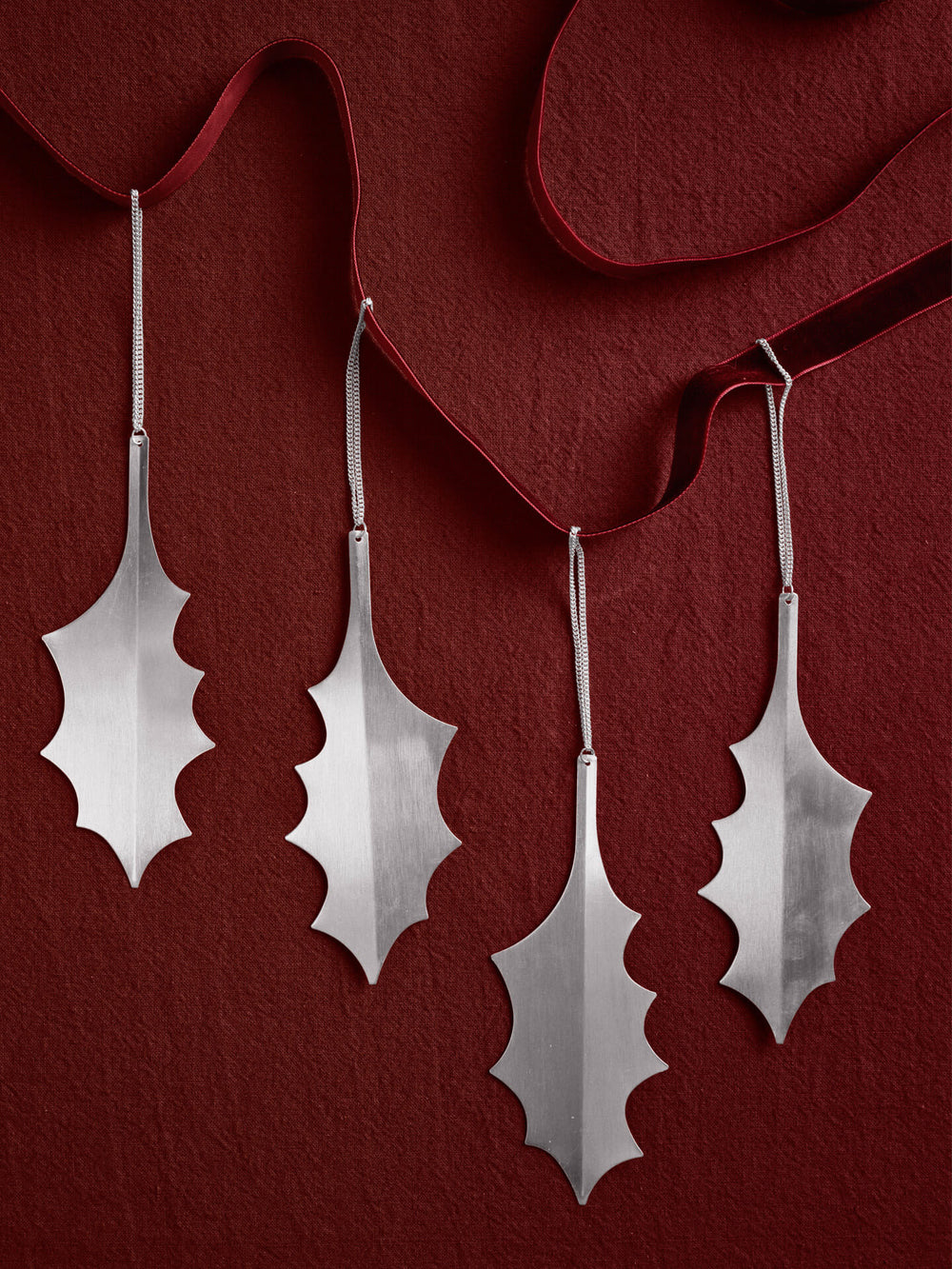 Four silver holly leaf ornaments on hanging chains, laid on deep red fabric with ribbon