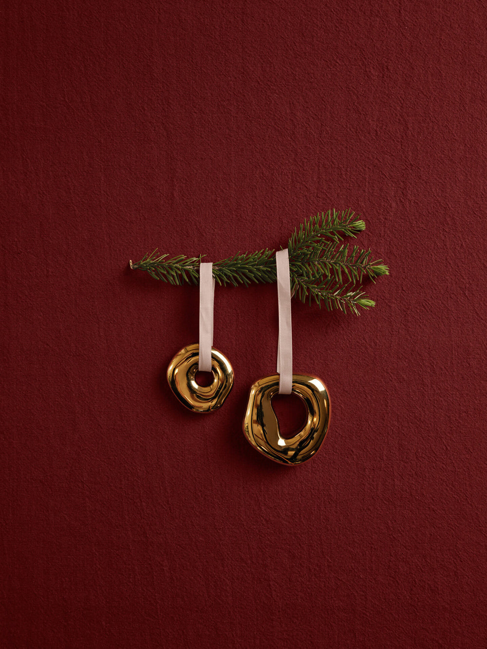 Two ring-shaped ornaments on pale ribbons, hung from a spruce sprig against a deep red backdrop