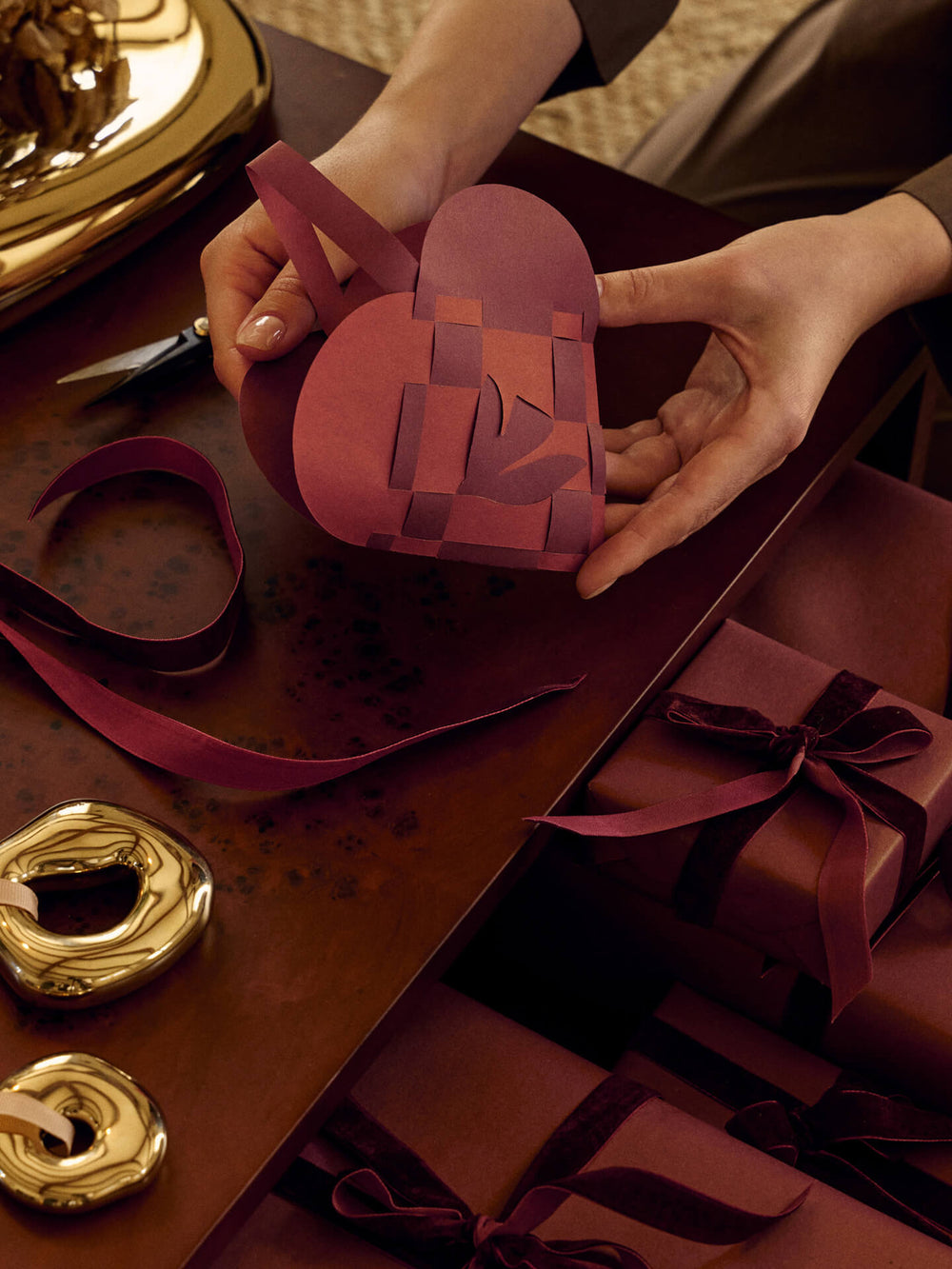 Hands weaving a red paper heart at a table with the brass ornaments, ribbon and wrapped gifts