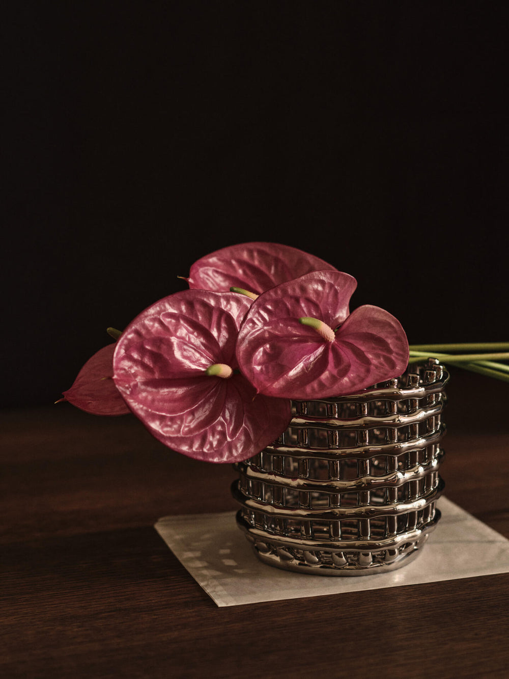 Woven-effect ceramic basket in silver on a dark table, holding pink anthurium flowers