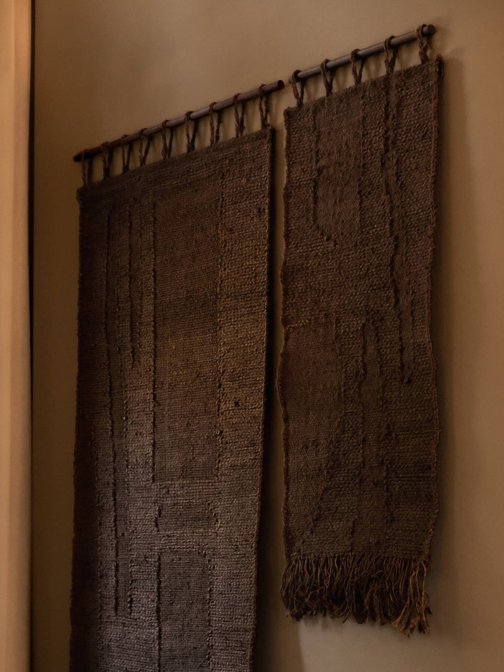 Two woven wall rugs in dark chocolate hanging side by side from bars on a beige wall