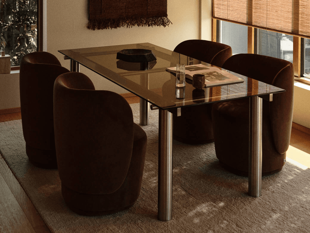 Dining table with tinted glass top in a sunlit room with four brown rounded armchairs and a wall hanging
