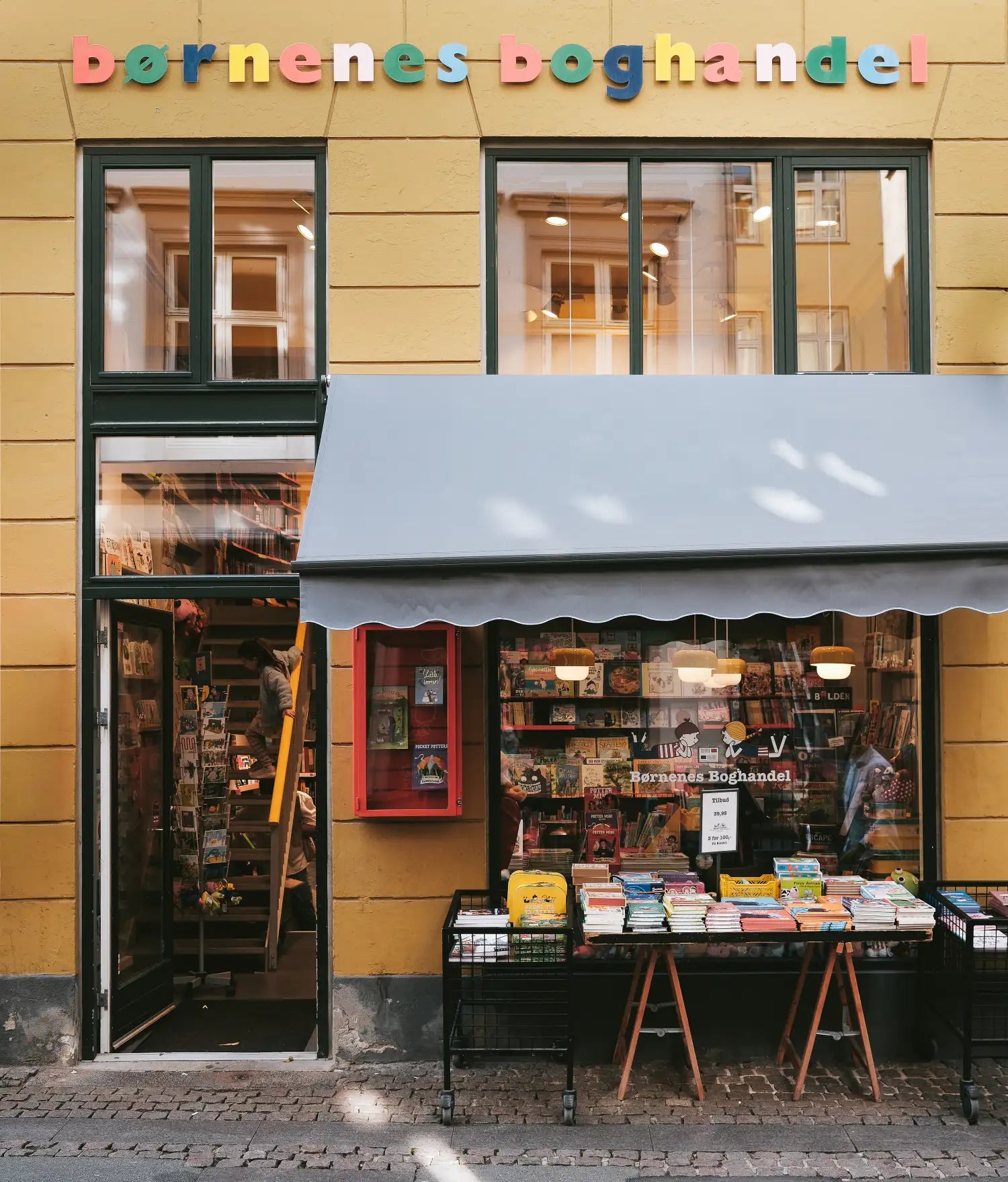 Børnenes Boghandel shopfront in yellow with blue awning, books on outdoor tables and a child on a ladder inside