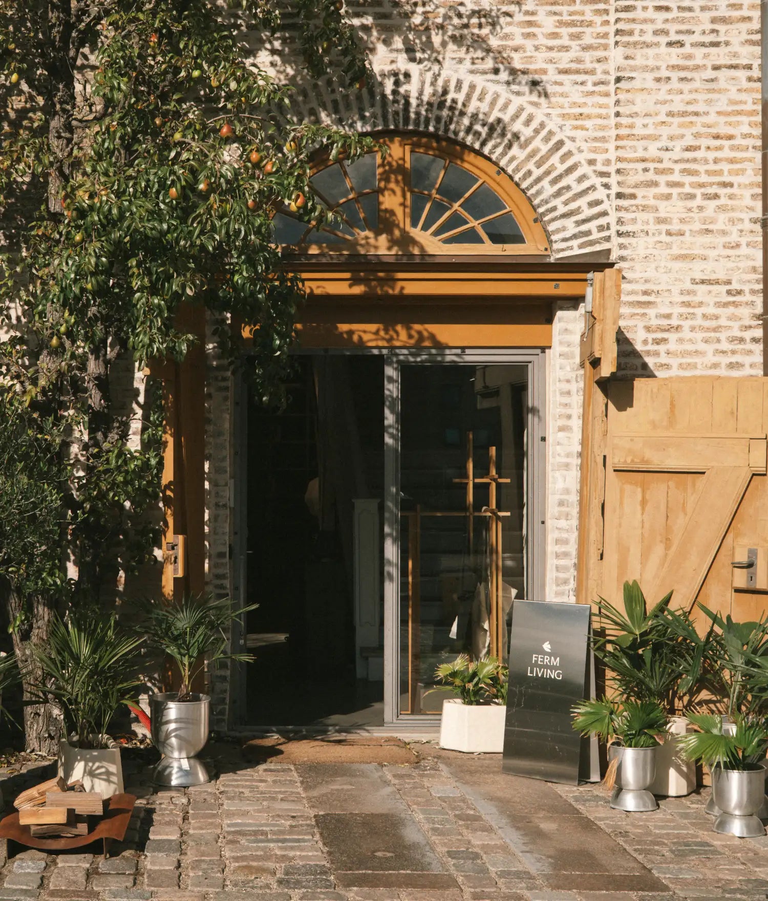 Open doorway in a brick building with an A-board sign, potted palms and a pear tree in a cobbled courtyard