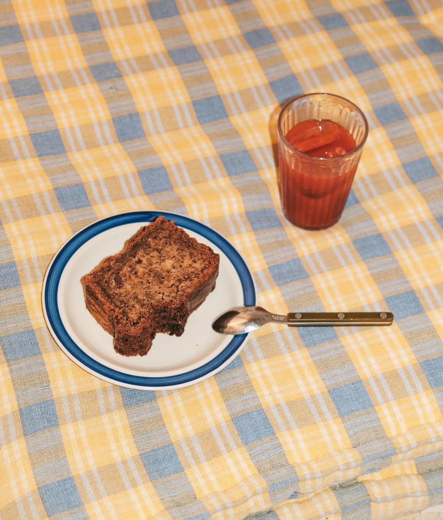 Yellow and blue checked tablecloth with a slice of cake on a blue rimmed plate, spoon and iced drink